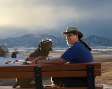 Ted and Fauna, his guide dog. They are both sitting on a bench looking over their shoulders at the camera. They are both wearing hats and a cloudy view of the rocky mountains can be seen in the background.