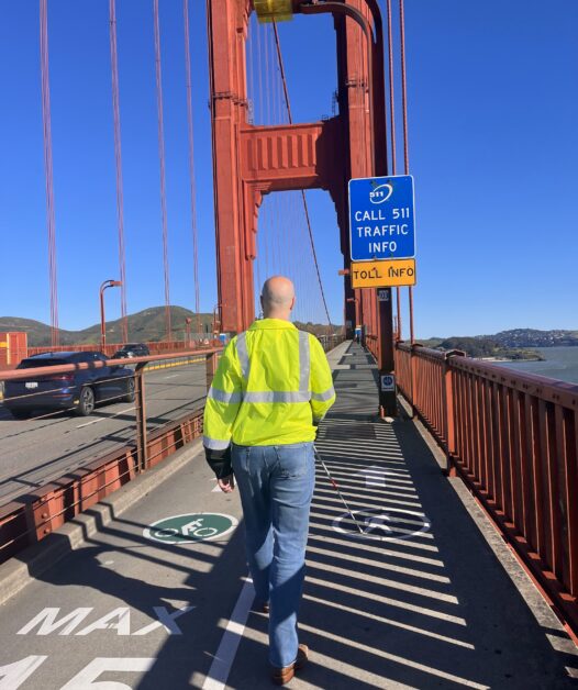 Chris Peterson walking on the Golden Gate Bridge and the large red metal support beams of the bridge in front of him.