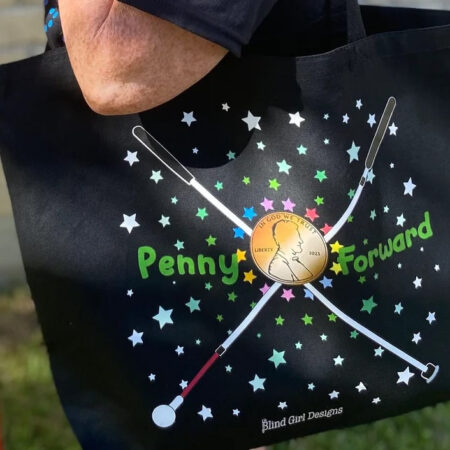 photo of Penny Forward black tote. The design has two white cane’s crossed in an x shape with a penny in the center. "Penny" is on the left of the penny with "Forward" on the right in green. There are colorful stars all around the graphic with Blind Girl Designs at the bottom.